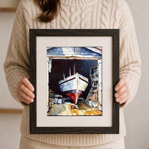 May include: A framed watercolor painting depicting a white boat with a red hull resting inside a dark wooden boathouse. The scene features a wooden floor, shelves with supplies, and a signature in the bottom right corner, all enclosed in a dark frame.
