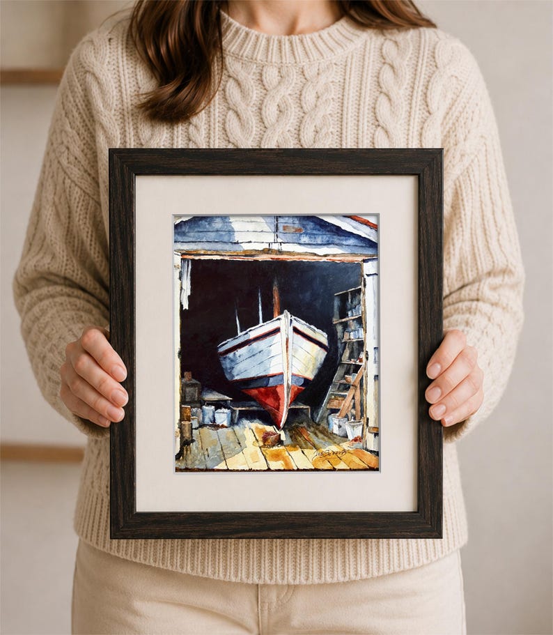 May include: A framed watercolor painting depicting a white boat with a red hull resting inside a dark wooden boathouse. The scene features a wooden floor, shelves with supplies, and a signature in the bottom right corner, all enclosed in a dark frame.