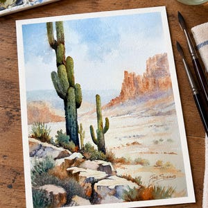 May include: A watercolor painting of a desert landscape featuring two tall saguaro cacti in the foreground, rocky terrain, and distant mesas under a light blue sky. The artist's signature, Joe Schaub, is visible in the bottom right corner.