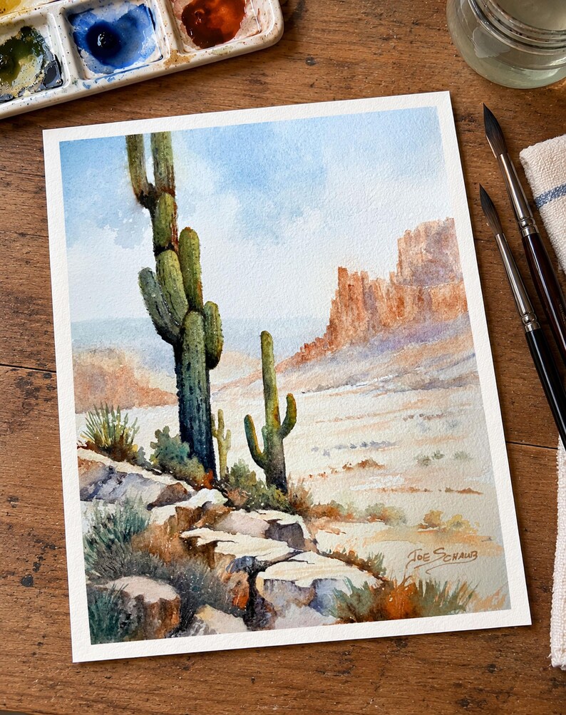 May include: A watercolor painting of a desert landscape featuring two tall saguaro cacti in the foreground, rocky terrain, and distant mesas under a light blue sky. The artist's signature, Joe Schaub, is visible in the bottom right corner.