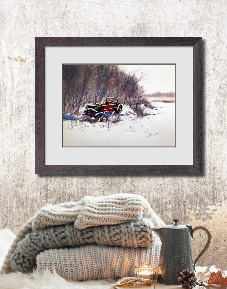 May include: A framed watercolour painting shows a vintage red truck in a snowy scene. The truck is partly covered in snow, set against bare trees and a muted sky. The dark brown frame surrounds the painting, which is displayed above a stack of grey blankets.