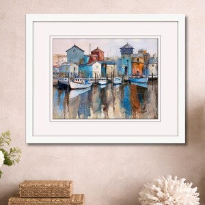 May include: Watercolor painting of a harbor scene with boats and buildings in shades of blue, red, and yellow. The artwork is framed in a white mat and frame. The water reflects the colors of the buildings and boats. The painting is displayed on a wooden surface.