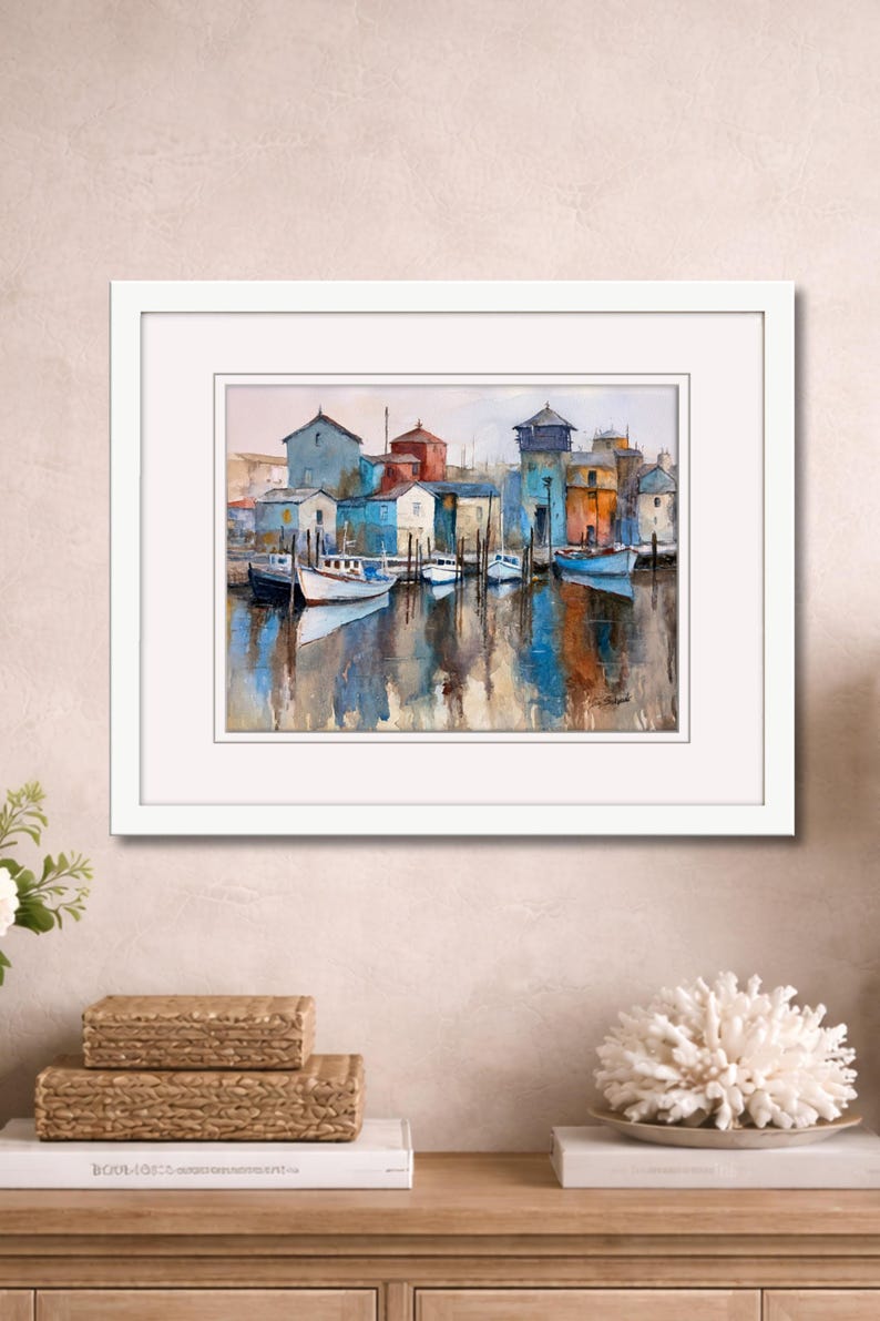 May include: Watercolor painting of a harbor scene with boats and buildings in shades of blue, red, and yellow. The artwork is framed in a white mat and frame. The water reflects the colors of the buildings and boats. The painting is displayed on a wooden surface.