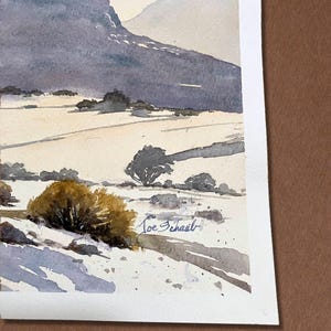 May include: Watercolor painting of a mountain landscape. The artwork features a large, dark purple mountain range in the background, with a field and small trees in the foreground. The artist's signature, "Joe Schail", is visible.