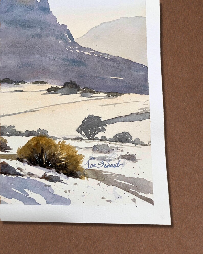 May include: Watercolor painting of a mountain landscape. The artwork features a large, dark purple mountain range in the background, with a field and small trees in the foreground. The artist's signature, "Joe Schail", is visible.