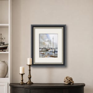 May include: A framed watercolor painting depicting several white fishing boats docked in shallow water under a cloudy sky. The artwork is enclosed in a dark gray frame with a white mat, showcasing a maritime scene with reflections on the water surface.