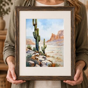 May include: A watercolor painting in a dark brown wooden frame depicting a desert landscape with two tall saguaro cacti, rocky terrain in the foreground, and distant mesas under a light blue sky. The artist's signature is visible in the corner.