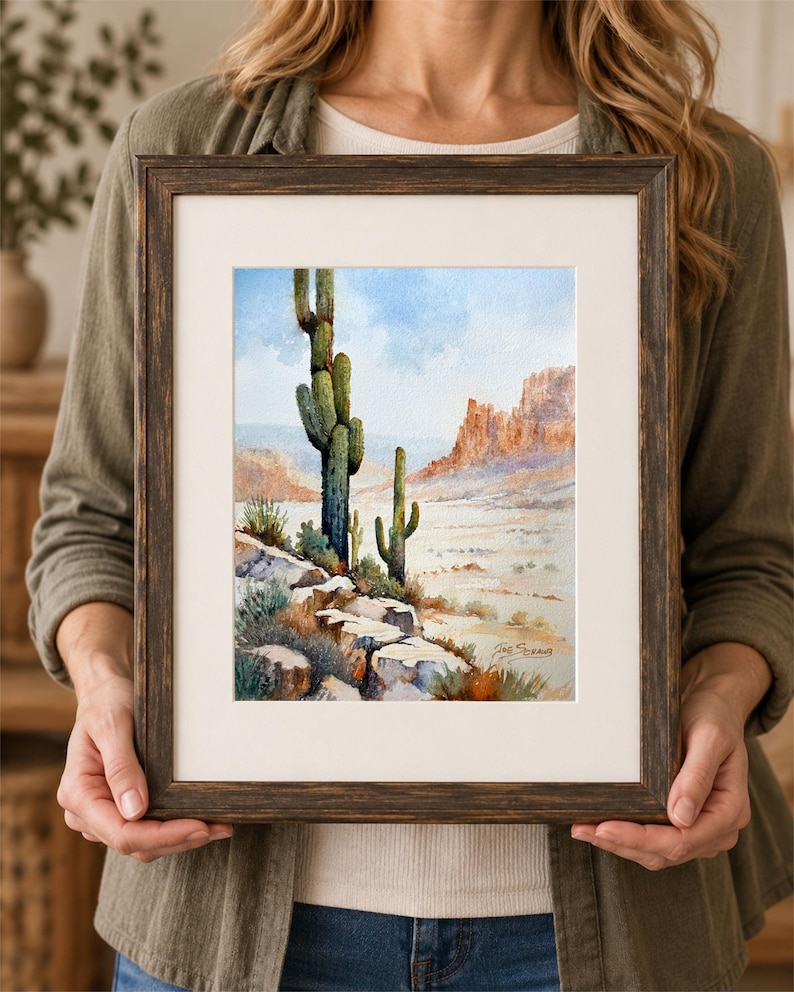May include: A watercolor painting in a dark brown wooden frame depicting a desert landscape with two tall saguaro cacti, rocky terrain in the foreground, and distant mesas under a light blue sky. The artist's signature is visible in the corner.