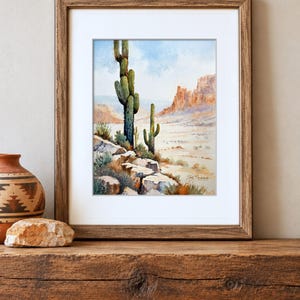 May include: A framed watercolor painting depicting a desert landscape with two saguaro cacti, rocky terrain, and distant mesas under a light blue sky. The artwork is housed in a brown wooden frame with a white mat and features a signature in the corner.
