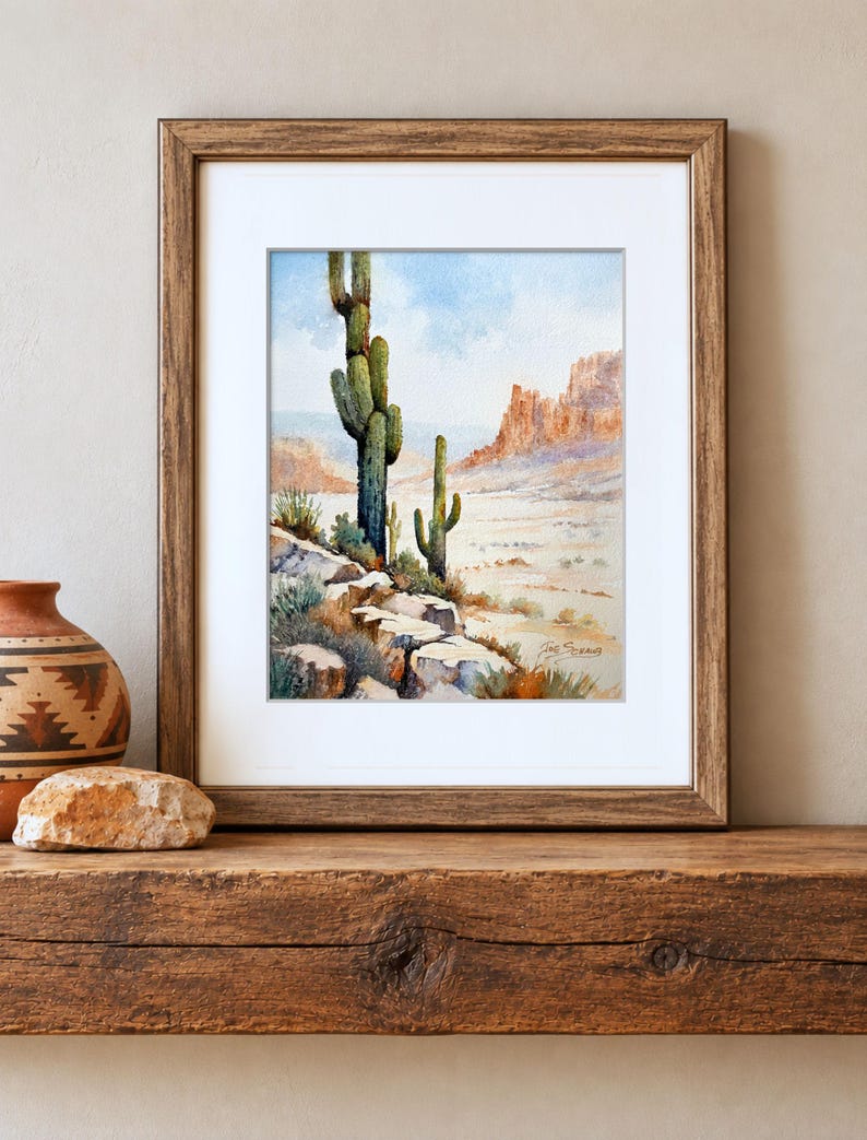May include: A framed watercolor painting depicting a desert landscape with two saguaro cacti, rocky terrain, and distant mesas under a light blue sky. The artwork is housed in a brown wooden frame with a white mat and features a signature in the corner.