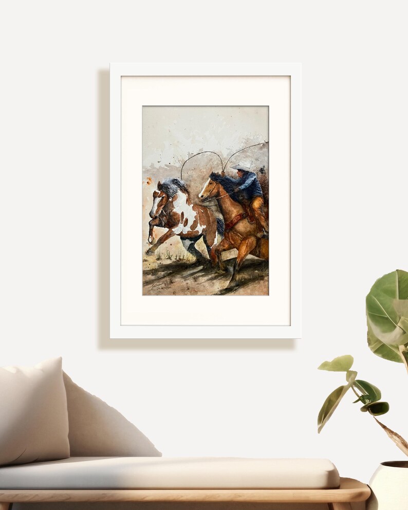 May include: Watercolor painting of two horses with riders in a white frame. One horse is brown and white, the other is brown. The riders are wearing cowboy hats and blue shirts. The artwork is displayed on a white wall.