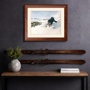 May include: A framed print of a skier on a snowy mountain slope, above a wooden console table. The table has a black metal frame and holds a gray vase with greenery and a stack of white books. Two vintage skis are mounted on the wall.
