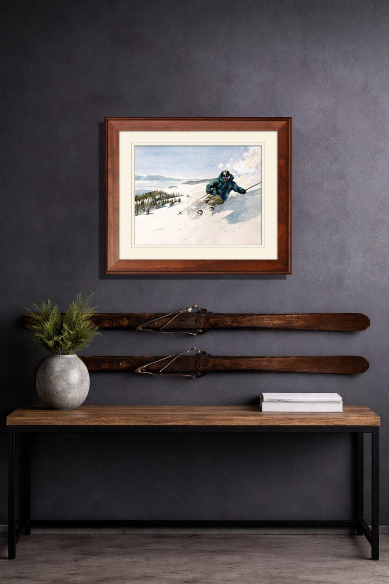 May include: A framed print of a skier on a snowy mountain slope, above a wooden console table. The table has a black metal frame and holds a gray vase with greenery and a stack of white books. Two vintage skis are mounted on the wall.