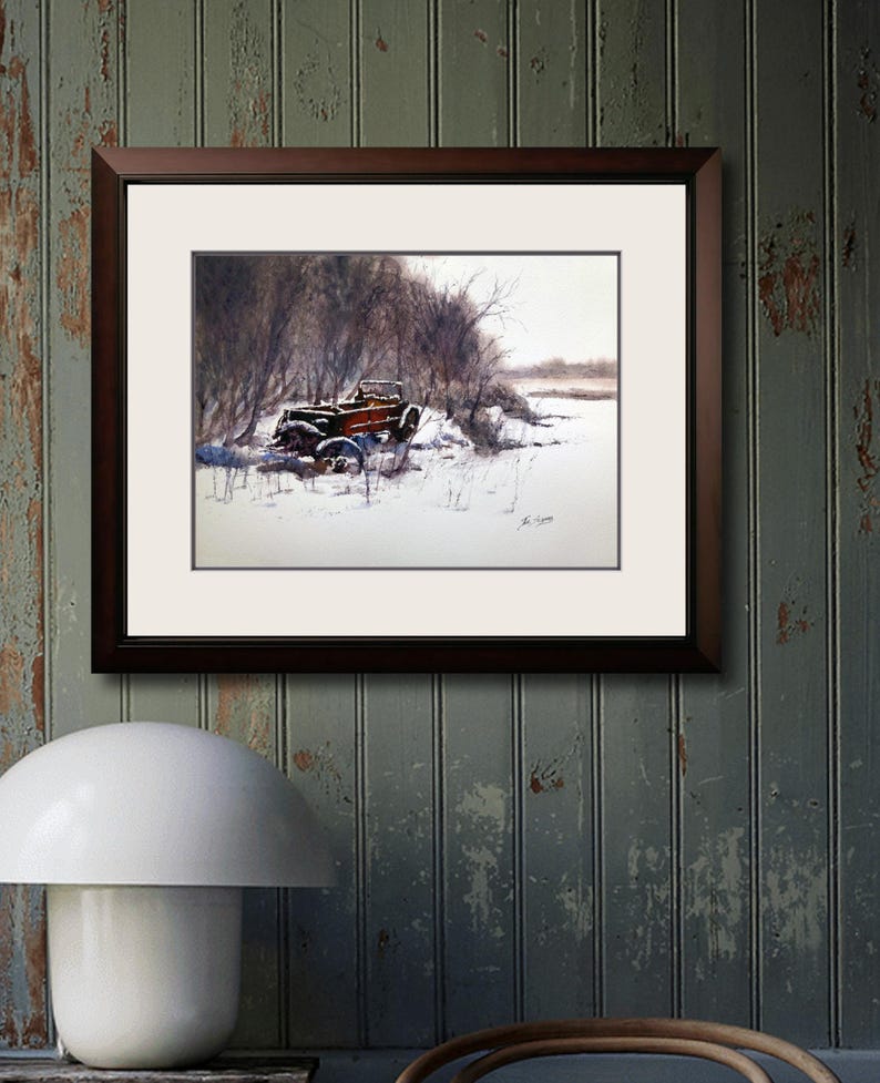 May include: A framed watercolour painting shows a vintage car in a snowy scene. The car, with a red body and black details, is partly covered in snow. The artwork is set against bare trees and a muted, wintery sky. The frame is dark brown.