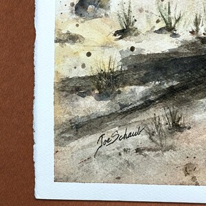 May include: Watercolor painting of a landscape scene with a neutral color palette. The artwork features a sandy ground, dark shadows, and tufts of grass. The artist's signature, "Joe Schaub," is visible in the lower right corner.