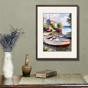 May include: A framed watercolor painting depicts several boats docked along a shoreline. The boats are painted in shades of red, white, and blue. Buildings line the shore under a blue sky. The painting is displayed on a wooden table with decorative items.