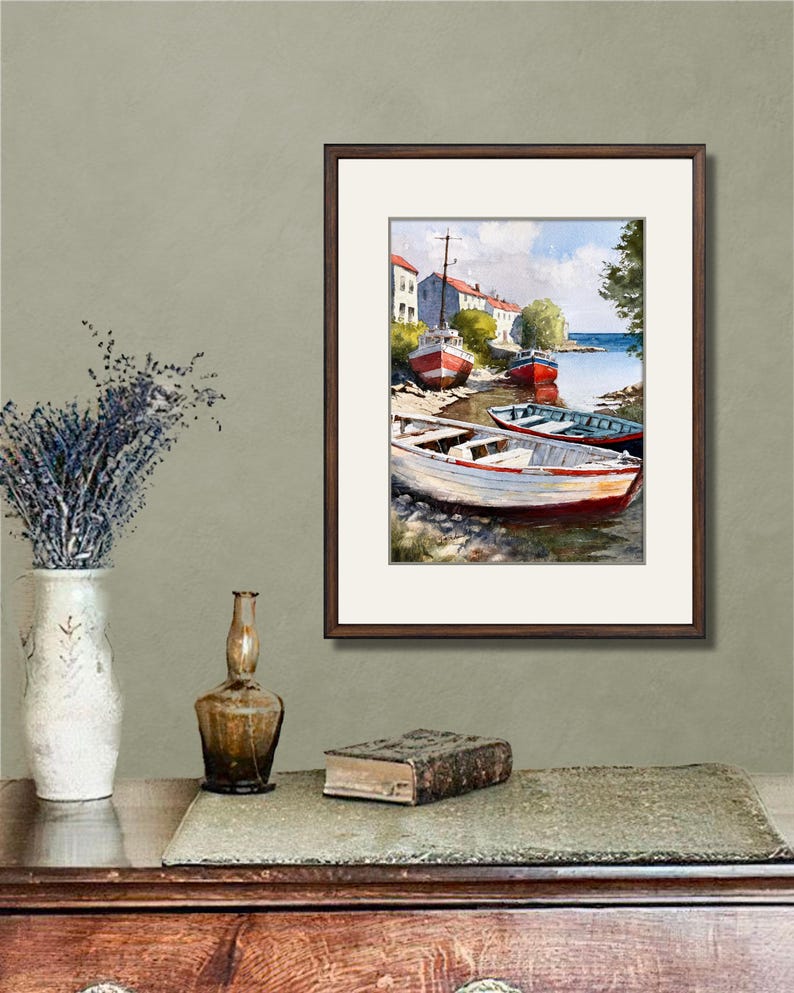 May include: A framed watercolor painting depicts several boats docked along a shoreline. The boats are painted in shades of red, white, and blue. Buildings line the shore under a blue sky. The painting is displayed on a wooden table with decorative items.