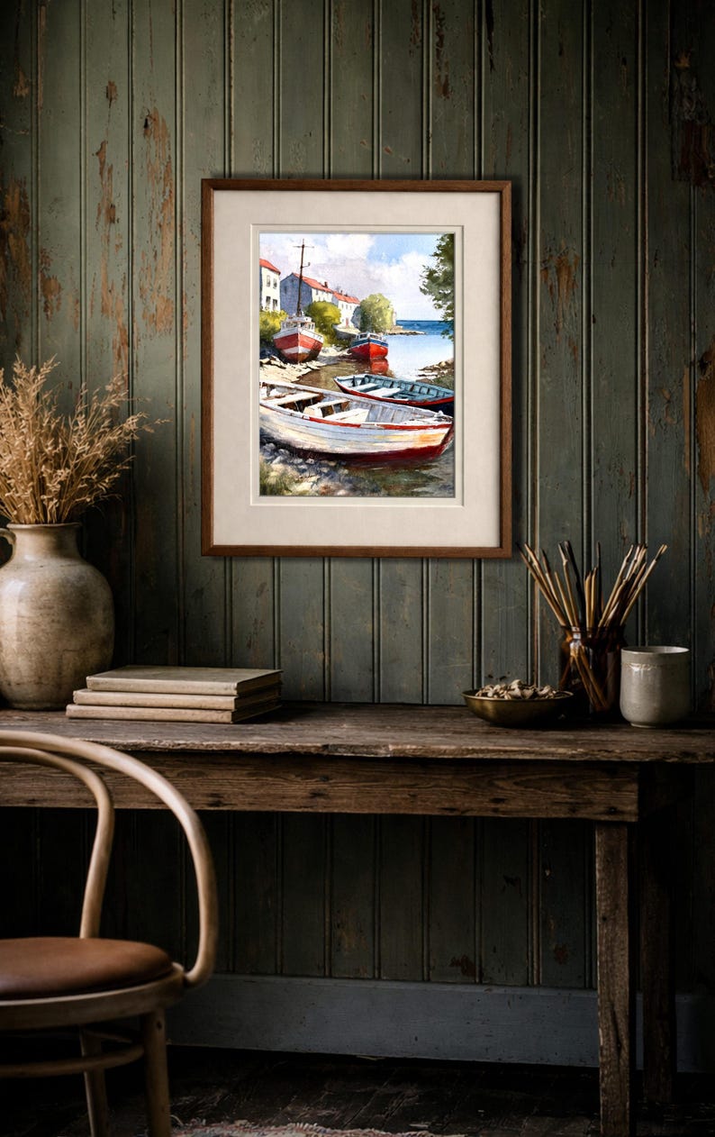 May include: A framed watercolor painting depicts a coastal scene with several small boats docked near a shore. The boats are painted in shades of red, white, and blue. The artwork is displayed on a wooden desk with other decorative items.