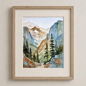 May include: Watercolor painting of a mountain landscape with a path. The artwork features a valley between mountains, with trees in the foreground and a light blue sky. The painting is framed in a light wood frame.