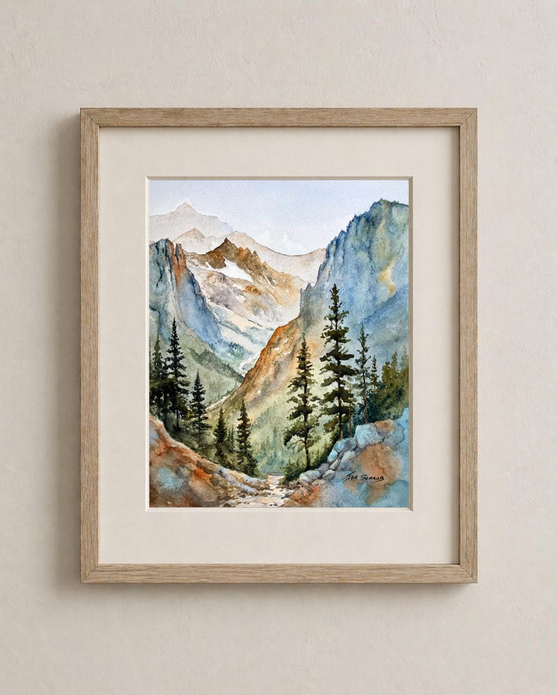 May include: Watercolor painting of a mountain landscape with a path. The artwork features a valley between mountains, with trees in the foreground and a light blue sky. The painting is framed in a light wood frame.