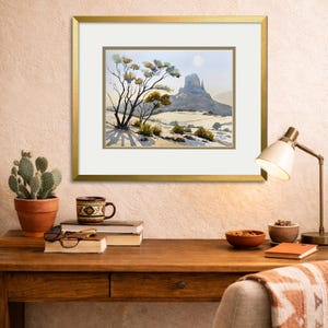 May include: A framed watercolor landscape painting depicting a desert scene with a large rock formation and sparse vegetation. The artwork is in a gold frame and is displayed on a wooden desk with a cactus, books, and a lamp.