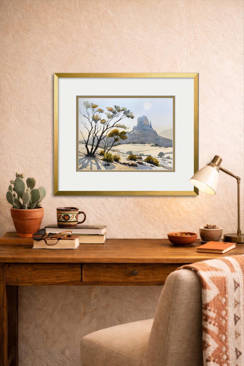 May include: A framed watercolor landscape painting depicting a desert scene with a large rock formation and sparse vegetation. The artwork is in a gold frame and is displayed on a wooden desk with a cactus, books, and a lamp.
