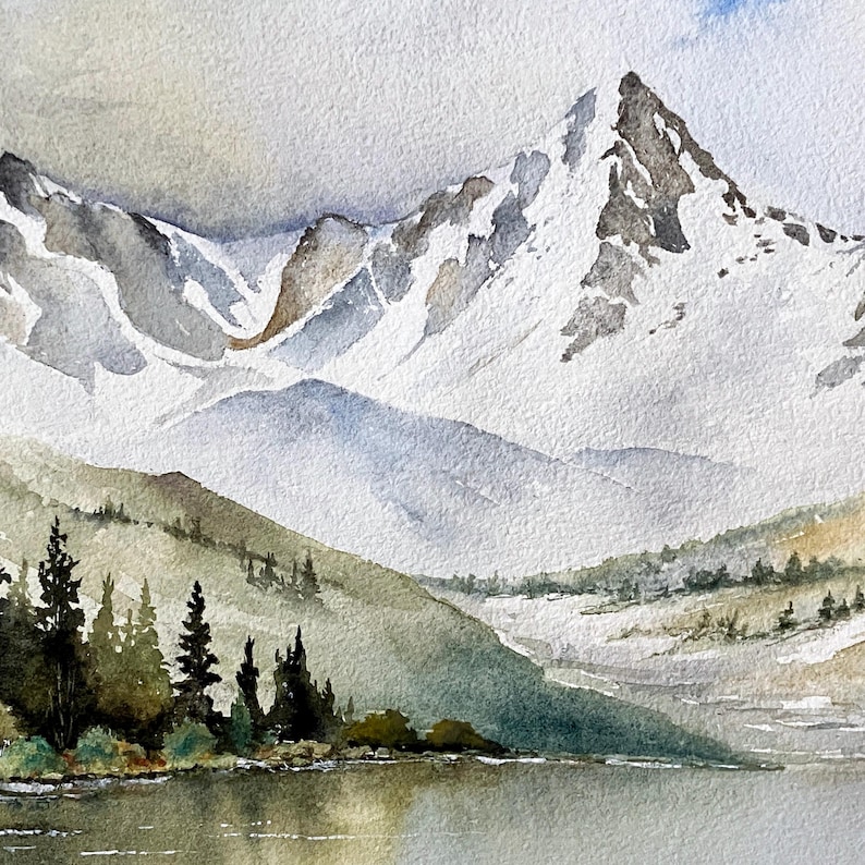 May include: A watercolor painting depicting a snow-capped mountain range rising above a calm lake. The foreground features dark green pine trees along the shoreline, while the peaks are rendered in shades of white, gray, and soft blue tones.