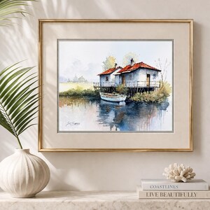 May include: Watercolor painting of two small houses on stilts over water, with a small boat. The houses have red roofs and white walls. The artwork is framed in a gold frame and is displayed on a white shelf.