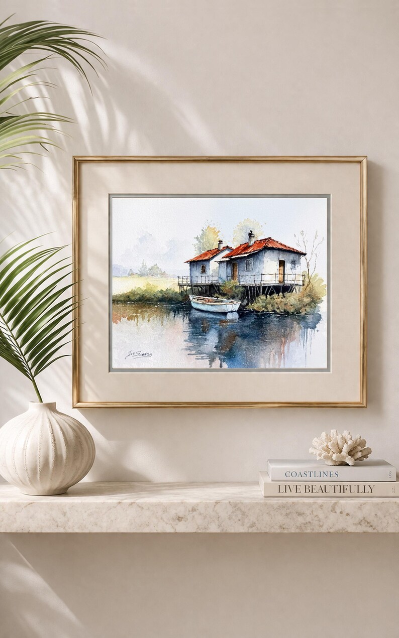 May include: Watercolor painting of two small houses on stilts over water, with a small boat. The houses have red roofs and white walls. The artwork is framed in a gold frame and is displayed on a white shelf.