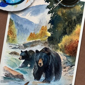 以下が含まれることがあります： 川の中にいる2匹の黒い熊を描いた水彩画。背景には山々が描かれています。青、緑、茶色を基調とした作品で、筆とパレットが描かれています。Joe Schueltのサイン入りです。