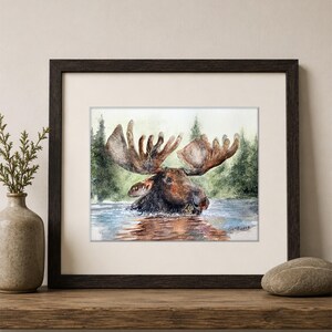 May include: A framed watercolor painting depicting a brown moose wading in water with a forest background. The artwork features a dark brown wooden frame, a white mat, and a signature in the bottom right corner of the painting, displayed on a shelf.