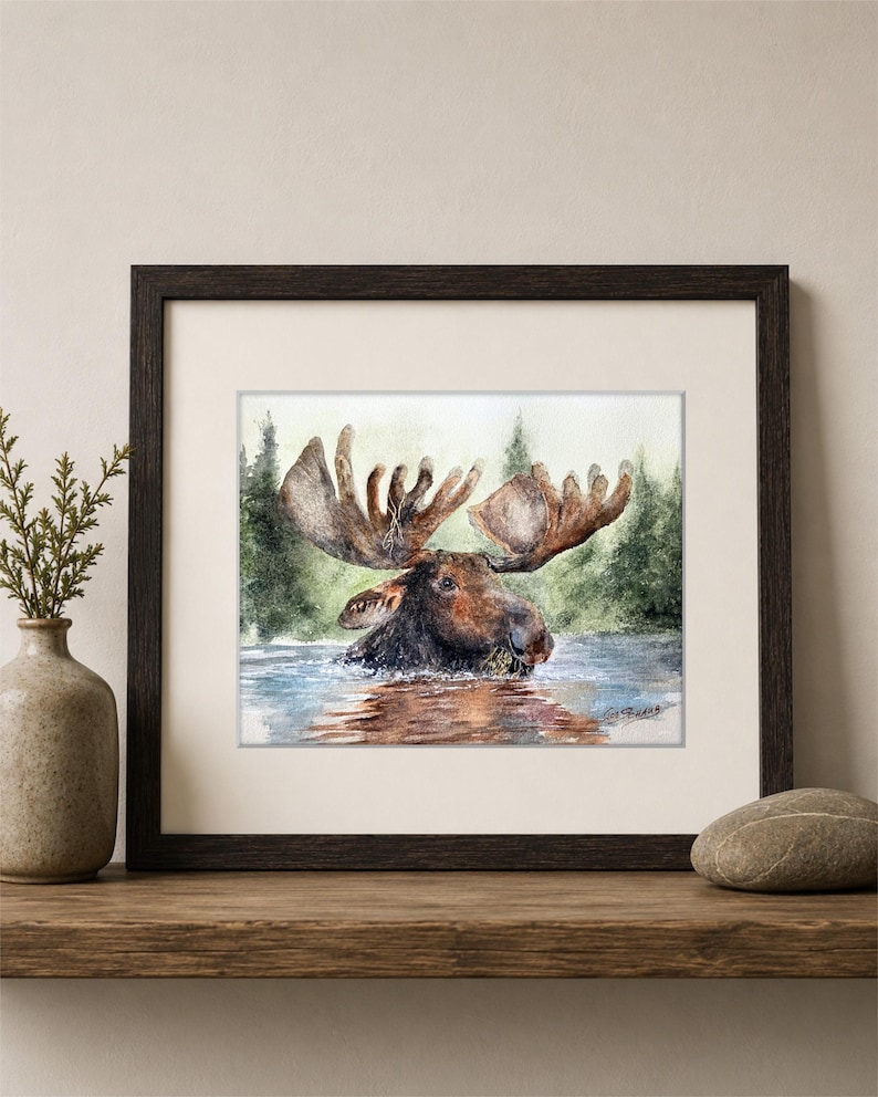 May include: A framed watercolor painting depicting a brown moose wading in water with a forest background. The artwork features a dark brown wooden frame, a white mat, and a signature in the bottom right corner of the painting, displayed on a shelf.