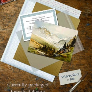 Puede incluir: Una pintura de paisaje en acuarela de monta&ntilde;as y &aacute;rboles con la marca de agua Sample, acompa&ntilde;ada de un certificado de autenticidad, una tarjeta de visita con el texto Watercolors By Joe, un pincel y un set de acuarelas en su embalaje.