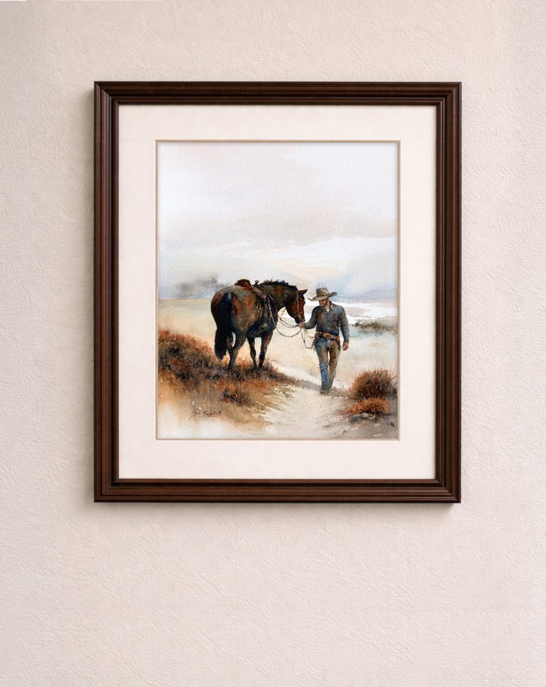 May include: Watercolor painting in a dark brown frame. The artwork depicts a cowboy walking alongside a horse on a dirt path. The horse is saddled and the cowboy is wearing a hat and jeans. The background shows a hazy landscape.