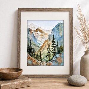 May include: Watercolor painting of a mountain landscape in a wooden frame. The artwork depicts a valley with tall evergreen trees, rocky cliffs, and snow-capped mountains in the background. The color palette includes blues, greens, and browns.