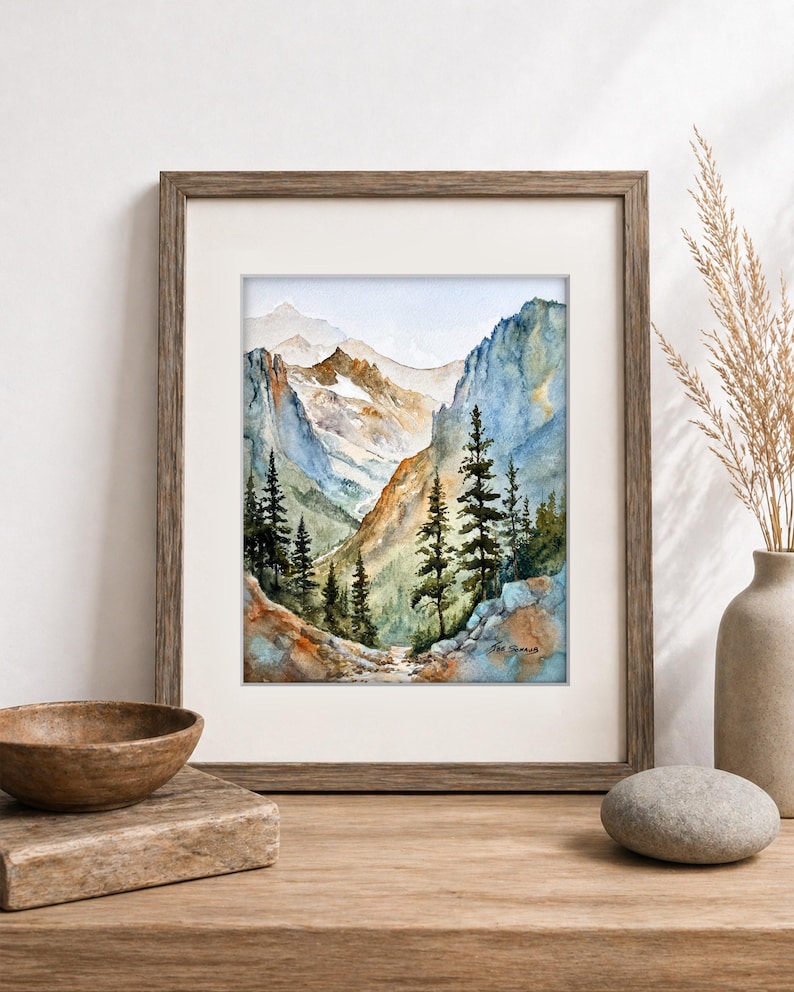 May include: Watercolor painting of a mountain landscape in a wooden frame. The artwork depicts a valley with tall evergreen trees, rocky cliffs, and snow-capped mountains in the background. The color palette includes blues, greens, and browns.