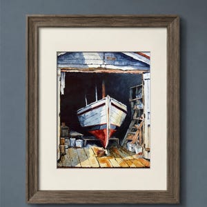 May include: A framed watercolor painting depicting a white boat with a red hull resting inside a dark wooden boathouse. The scene features a wooden floor, storage shelves, and a signature in the bottom right corner. The frame is dark brown wood.