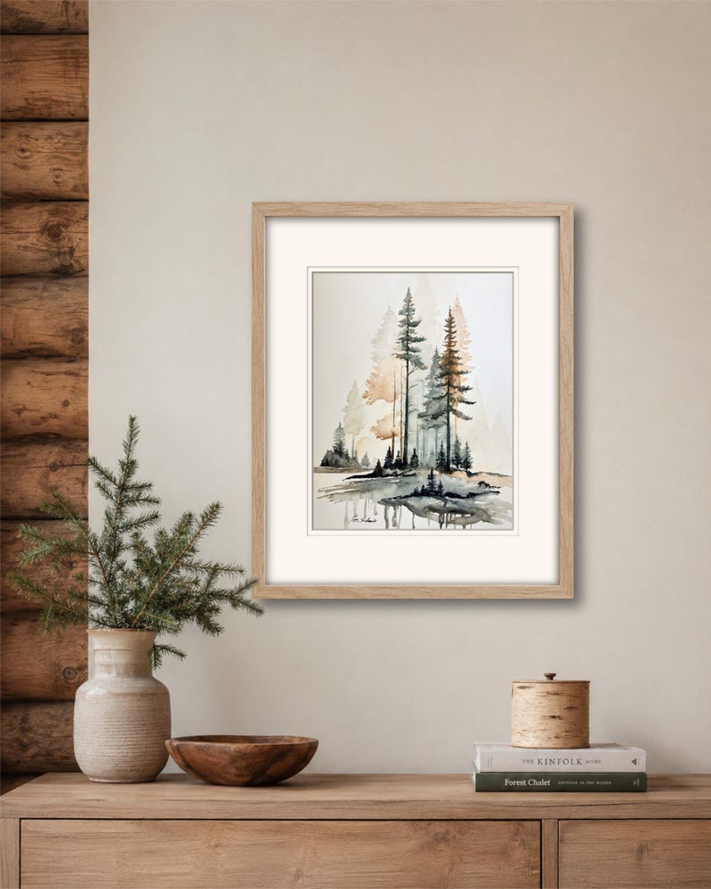 May include: A framed watercolor painting depicting a forest scene with tall pine trees in shades of green, brown, and muted orange, reflected in a calm body of water. The artwork is enclosed in a light-toned wooden frame with a white mat border.