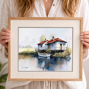 May include: Watercolor painting of two white houses with red roofs on stilts, next to a calm body of water. A small white boat is docked nearby. The artwork is framed in a light brown wooden frame.