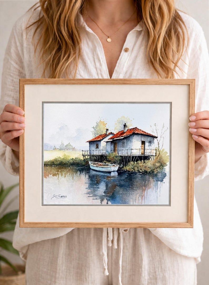 May include: Watercolor painting of two white houses with red roofs on stilts, next to a calm body of water. A small white boat is docked nearby. The artwork is framed in a light brown wooden frame.