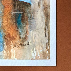 May include: Watercolor painting of a building with a blue sky. The artwork features shades of brown, orange, and blue. The artist's signature, "Joe Schaut," is visible in the lower left corner. The painting is on white paper.