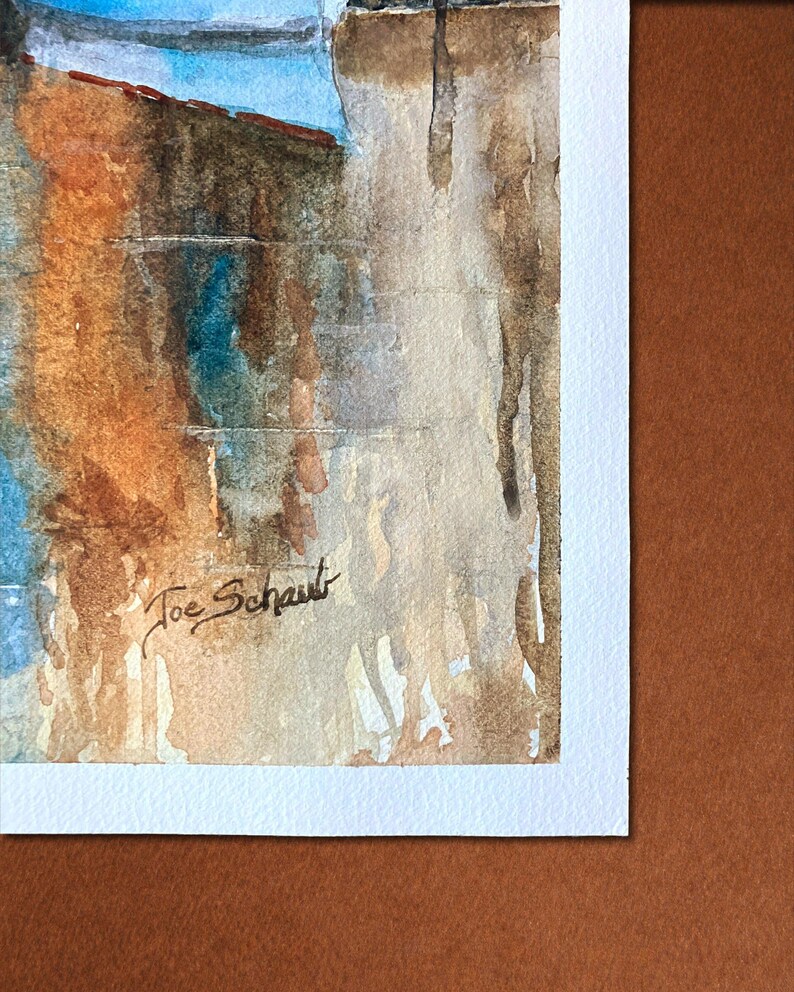 May include: Watercolor painting of a building with a blue sky. The artwork features shades of brown, orange, and blue. The artist's signature, "Joe Schaut," is visible in the lower left corner. The painting is on white paper.