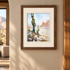 May include: A framed watercolor painting depicting a desert landscape with two saguaro cacti in the foreground, rocky terrain, and distant mesas under a light blue sky. The artwork is enclosed in a dark brown wooden frame with a cream mat.