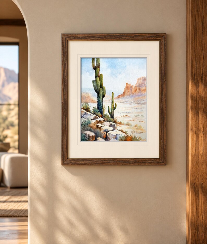 May include: A framed watercolor painting depicting a desert landscape with two saguaro cacti in the foreground, rocky terrain, and distant mesas under a light blue sky. The artwork is enclosed in a dark brown wooden frame with a cream mat.