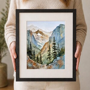 May include: A framed watercolor painting of a mountain landscape. The artwork features a path winding through a valley, with tall evergreen trees and towering mountains in the background. The painting is in a black frame with a white mat.