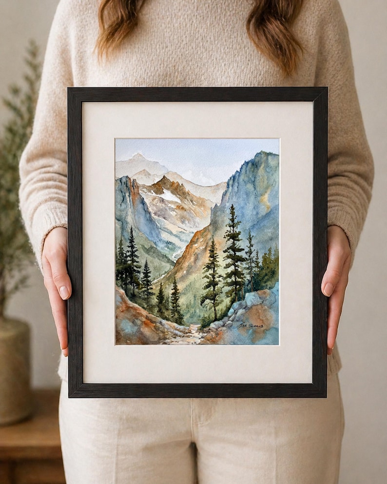 May include: A framed watercolor painting of a mountain landscape. The artwork features a path winding through a valley, with tall evergreen trees and towering mountains in the background. The painting is in a black frame with a white mat.