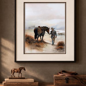 May include: Framed watercolor print depicting a cowboy leading a horse along a path. The artwork features muted tones of brown, beige, and blue, with a black frame and a cream-colored mat. A bronze horse figurine and a wooden box are on a wooden surface.