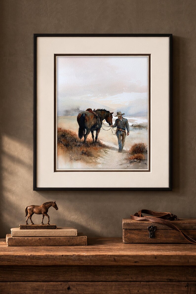 May include: Framed watercolor print depicting a cowboy leading a horse along a path. The artwork features muted tones of brown, beige, and blue, with a black frame and a cream-colored mat. A bronze horse figurine and a wooden box are on a wooden surface.