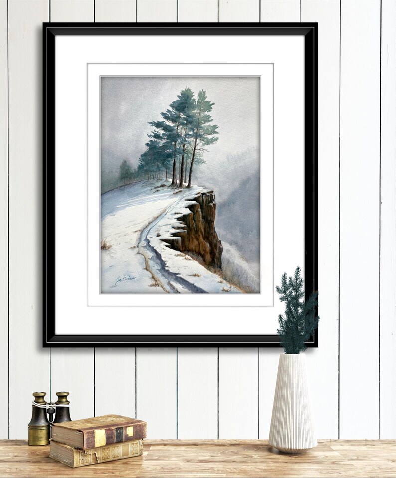 K&ouml;nnte beinhalten: Aquarellmalerei eines schneebedeckten Klippenrandes mit immergr&uuml;nen B&auml;umen. Das Kunstwerk ist in einem schwarzen Rahmen gerahmt und wei&szlig; mattiert. Das Gem&auml;lde zeigt eine Winterlandschaft mit einem Pfad entlang der Klippe.