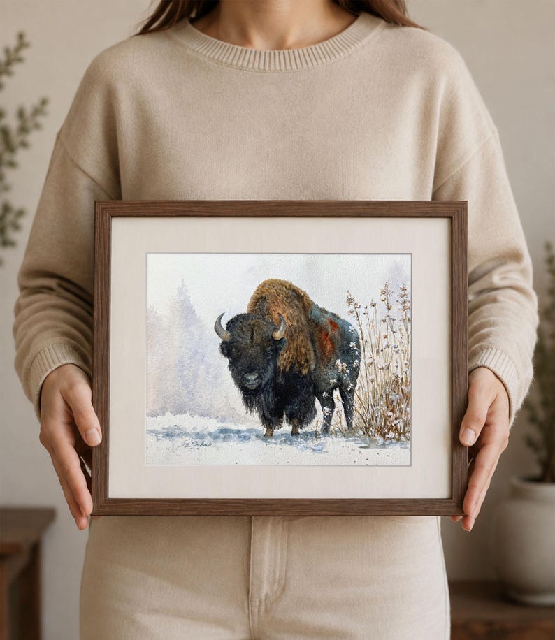 May include: A framed watercolor painting of a bison standing in a snowy landscape with dry grasses. The bison has dark brown and black fur with a lighter brown hump. The artwork is enclosed in a dark brown wooden frame with a white mat border.